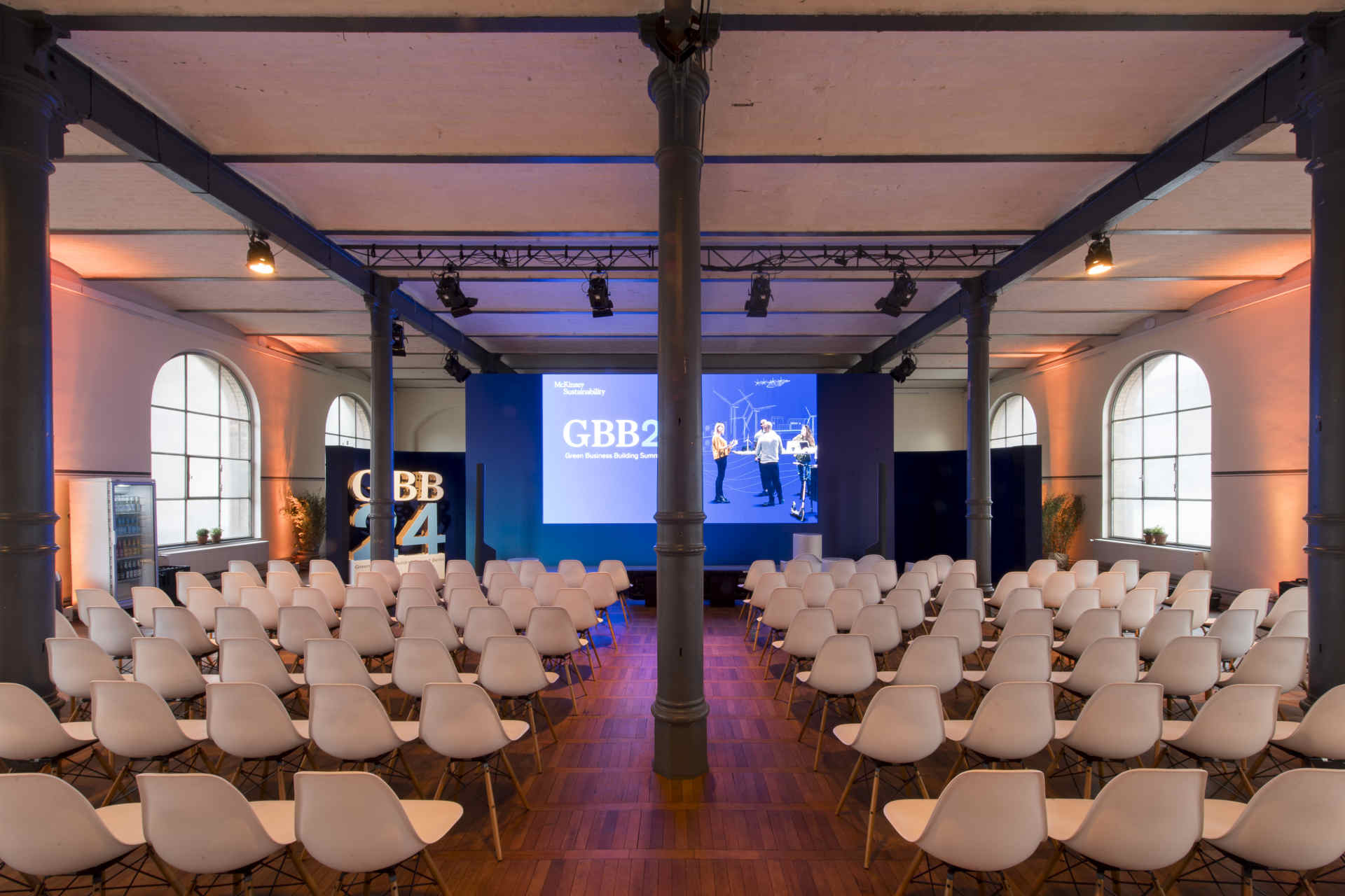 Saal mit Rundfenstern, weißer Reihenbestuhlung und Bühne mit Leinwand